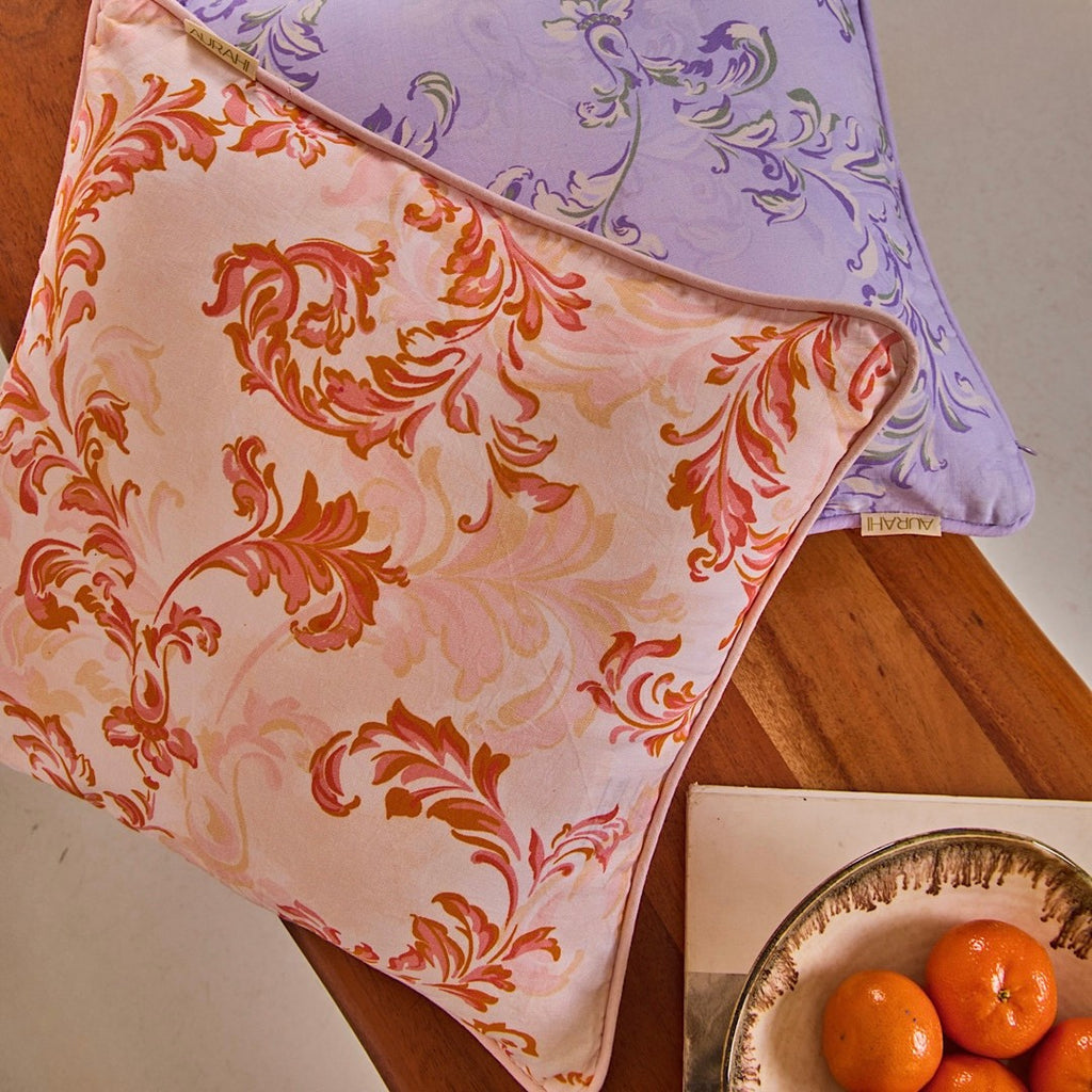 A top-down view of two stacked cushion covers made from hemp fabric, featuring an orange and pink damask-style scroll pattern on top and a lavender one underneath, styled on a wooden table next to a bowl of oranges.