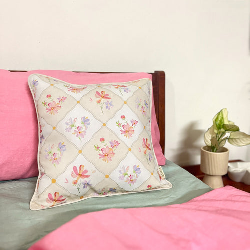 A lifestyle shot of a floral cushion cover made from hemp fabric, featuring a diamond lattice design with colorful watercolor wildflowers, styled on a bed with pink pillows and a light green sheet next to a small potted plant.