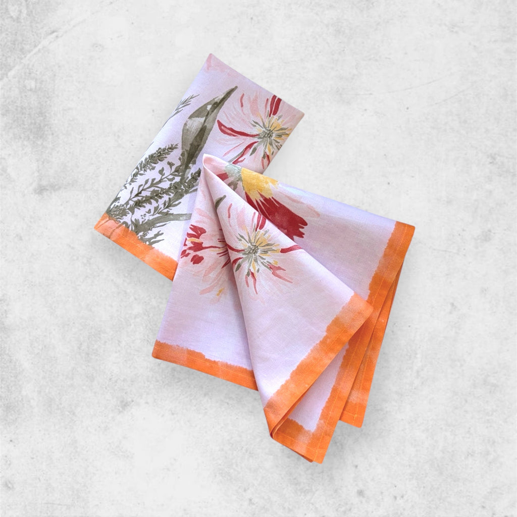 A folded sena table napkin featuring delicate hand-painted botanical artwork and a vibrant orange hand-painted border on a light pink base.